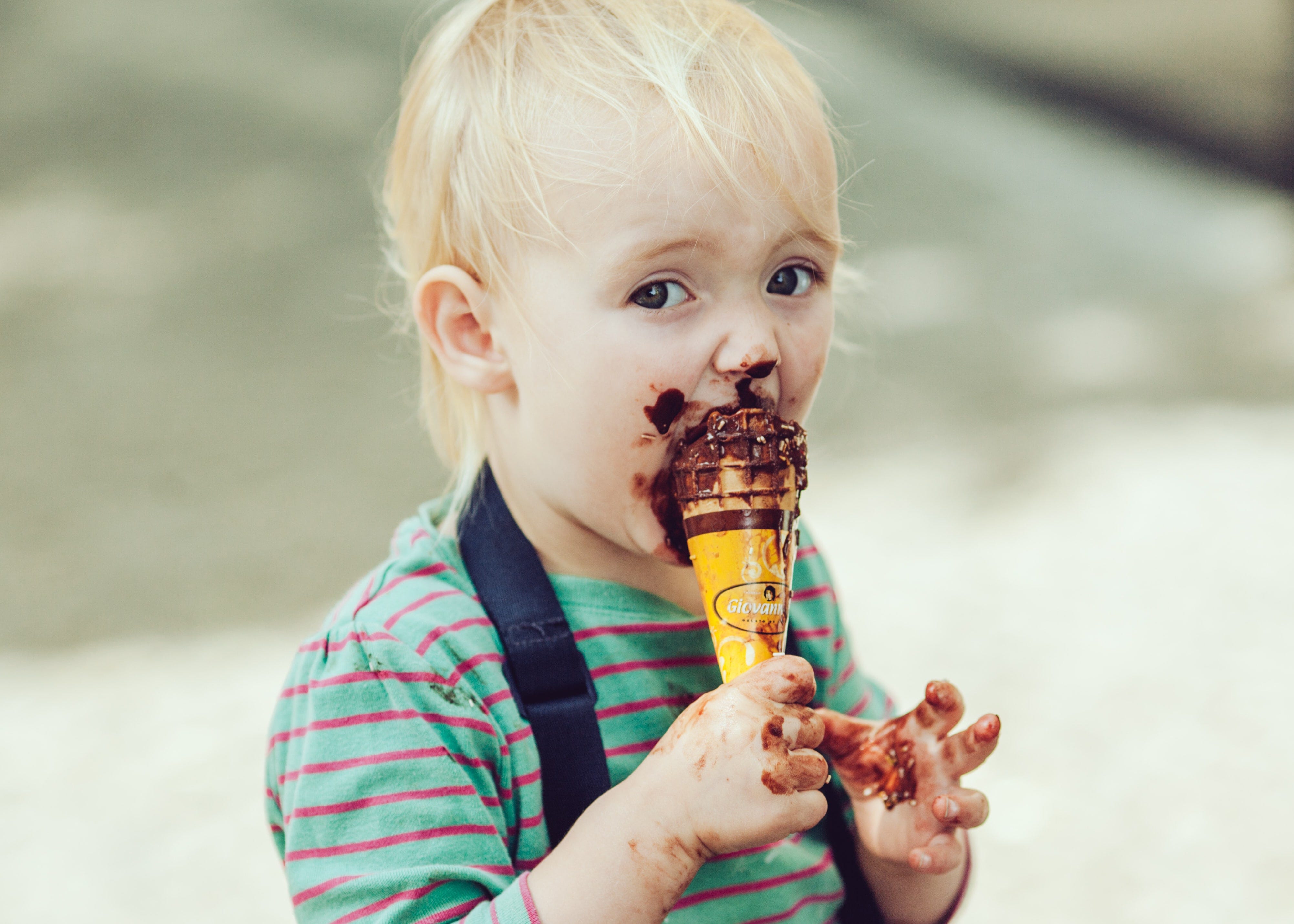 A young child outdoors eating chocolate icecream, with chocolate covering their face and hands