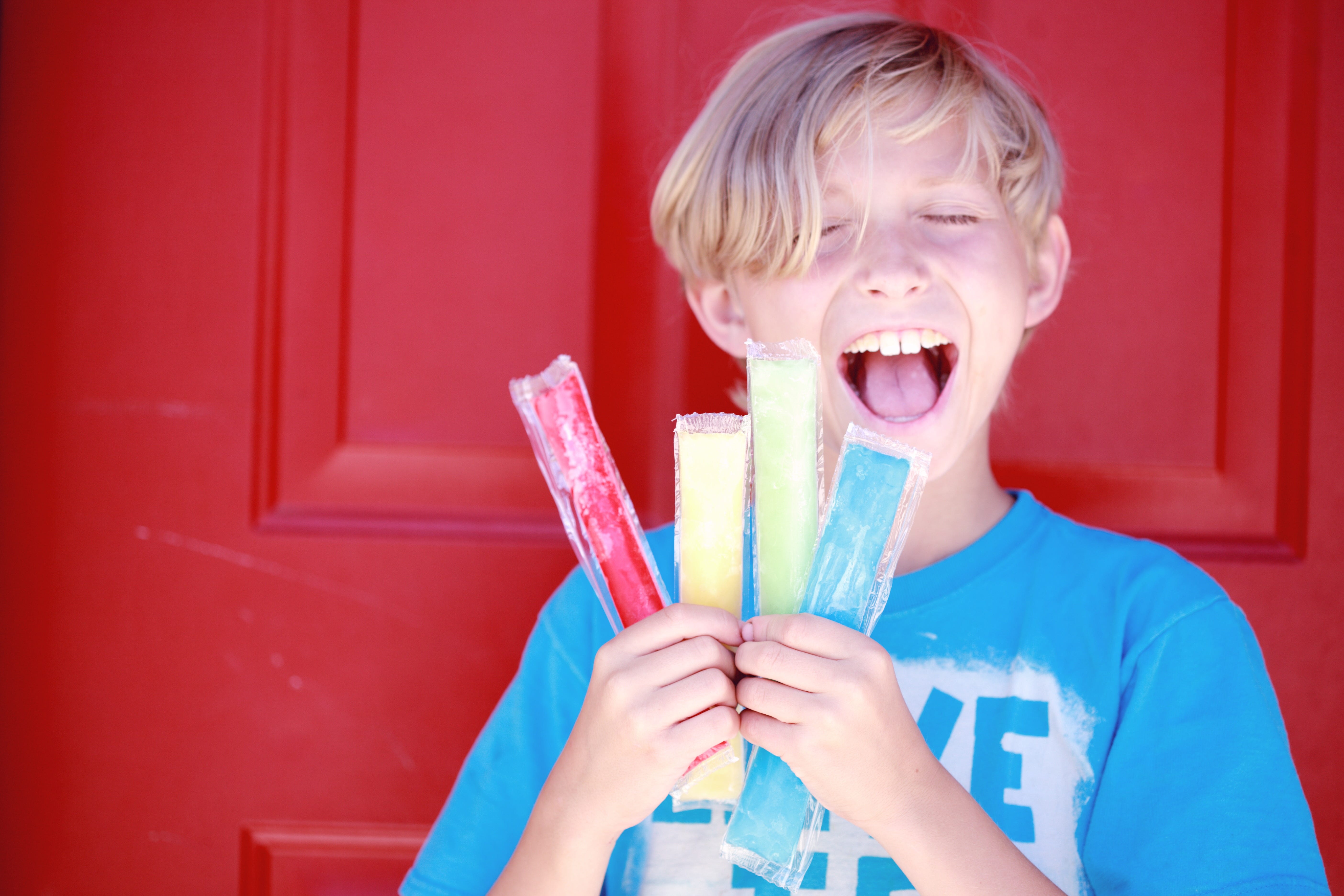 A child standing in front of a red door wearing a blue t-shirt, holding 4 ice pops and smiling 